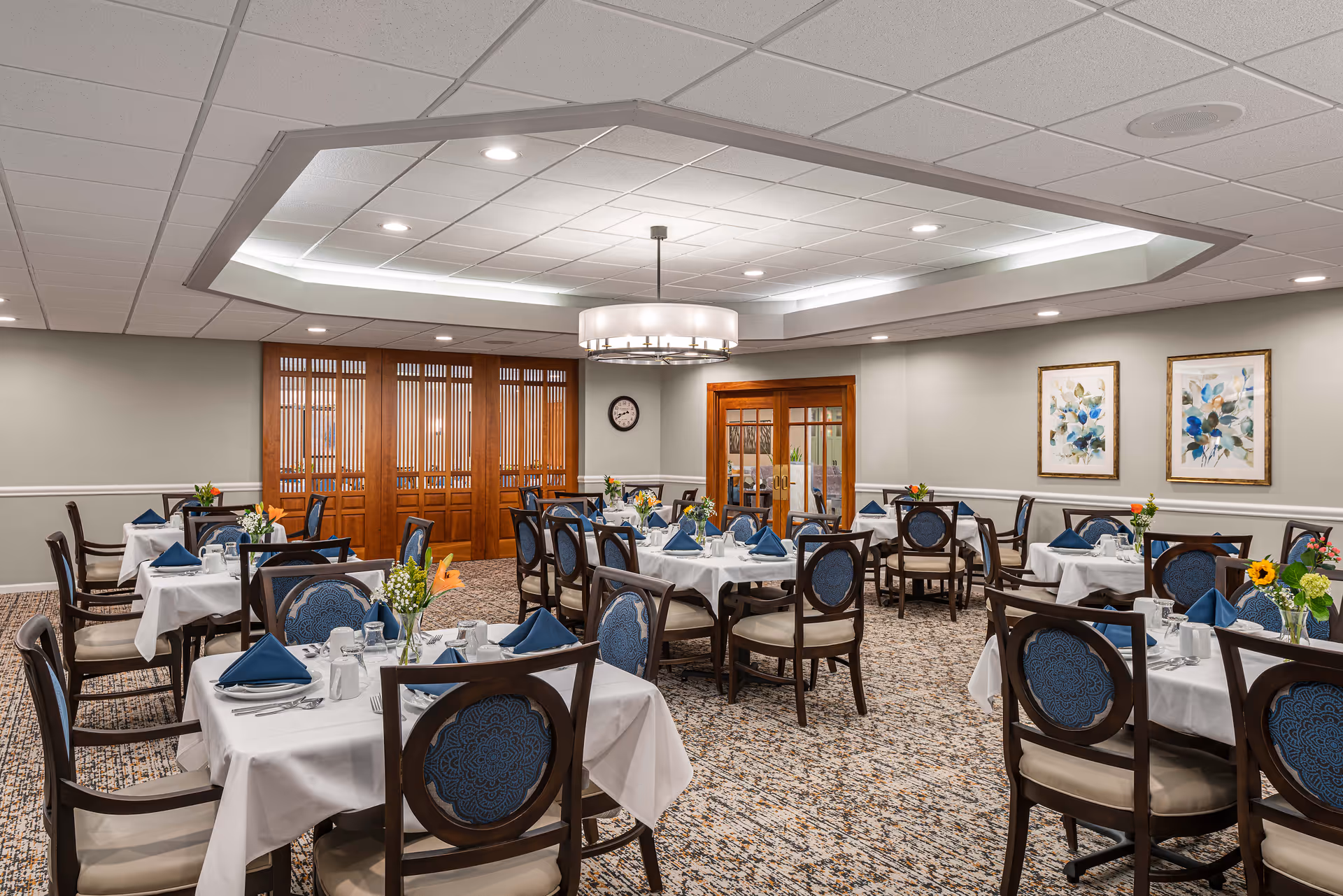 A well-lit dining room with multiple tables covered in white tablecloths, each set with blue napkins, plates, cups, and silverware. The chairs have dark wooden frames with blue patterned cushions. The room features a decorative ceiling light fixture, carpeted floor, wooden folding doors, and framed floral artwork on the walls.