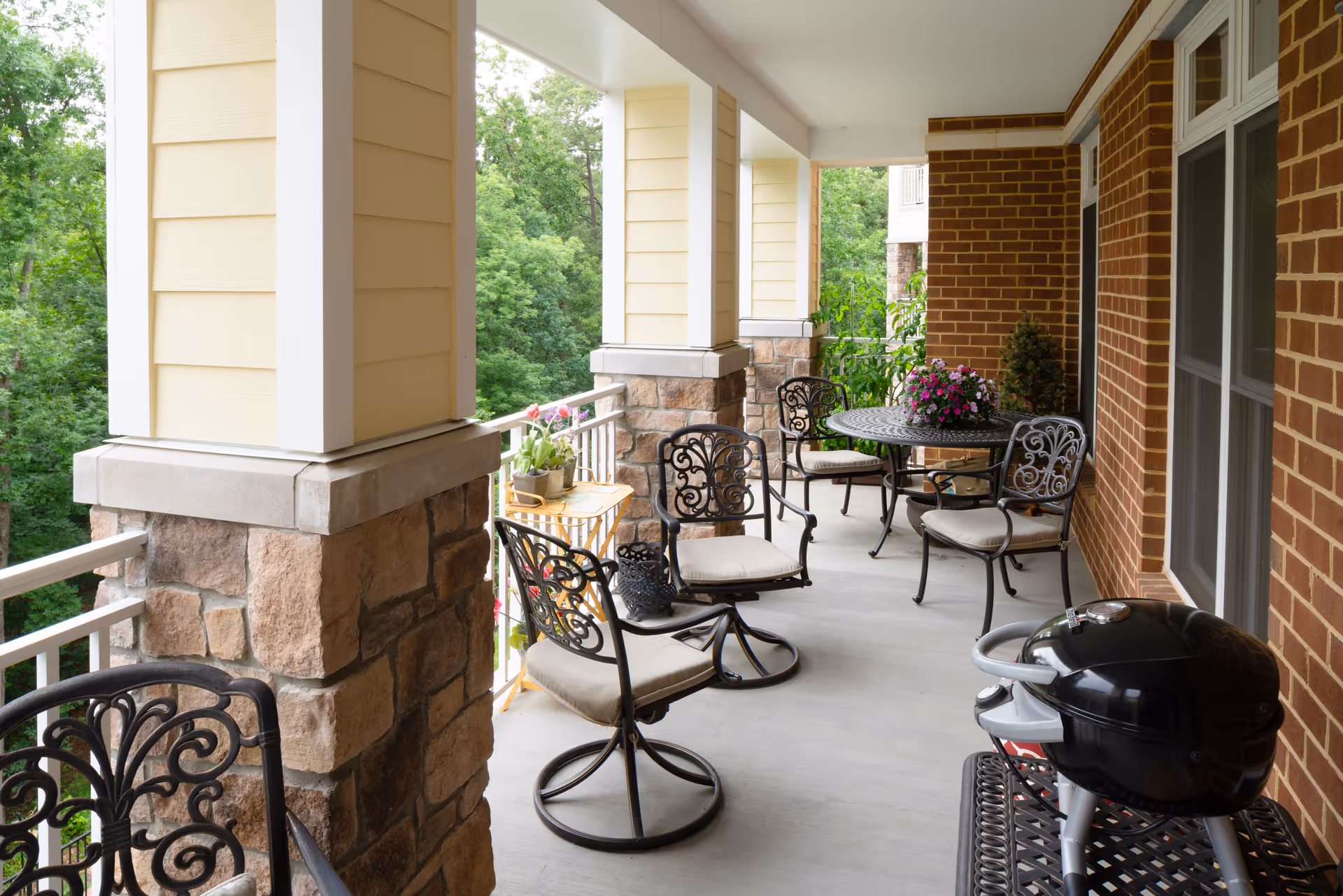 Covered outdoor patio area with cushioned wrought iron chairs and a round table with a flower arrangement. The patio has stone and brick pillars and overlooks a green wooded area. A small black grill is also visible on the right side.