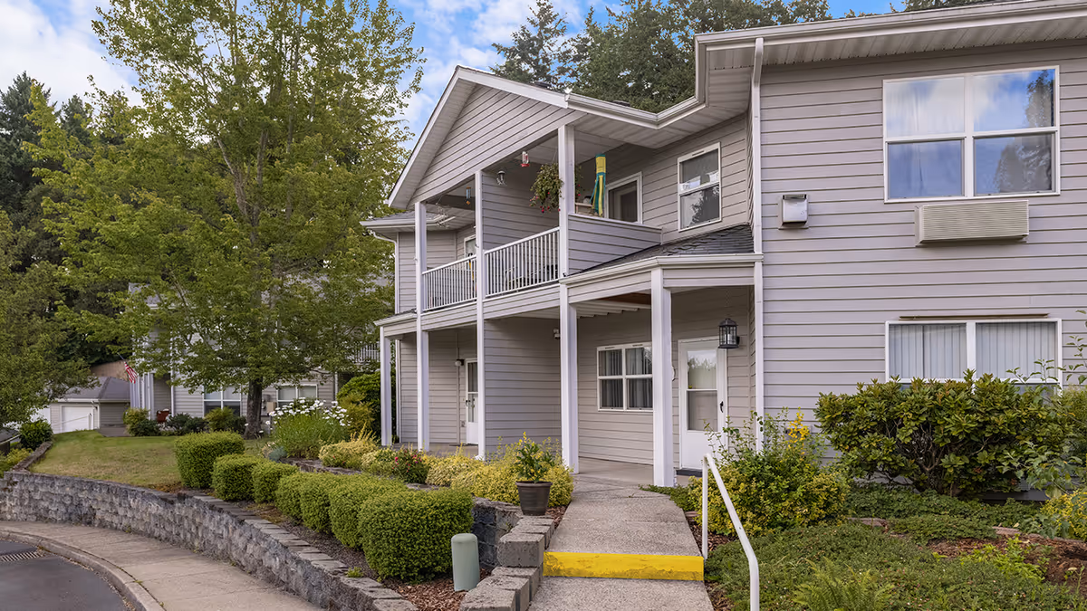 Two-story light-gray residential building with balconies, landscaped shrubs, and a walkway leading to the entrance.