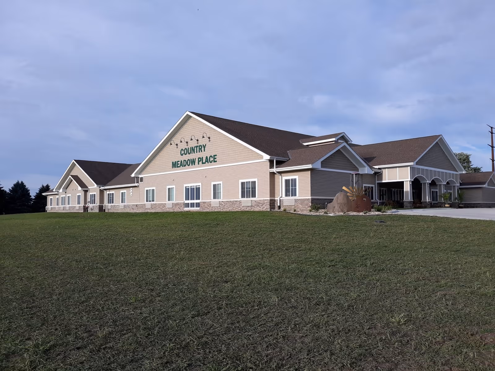 Exterior view of a single-story senior living facility named Country Meadow Place, featuring beige siding, stone accents along the lower walls, multiple windows, and a covered entrance. The building is surrounded by a large grassy area under a partly cloudy sky.