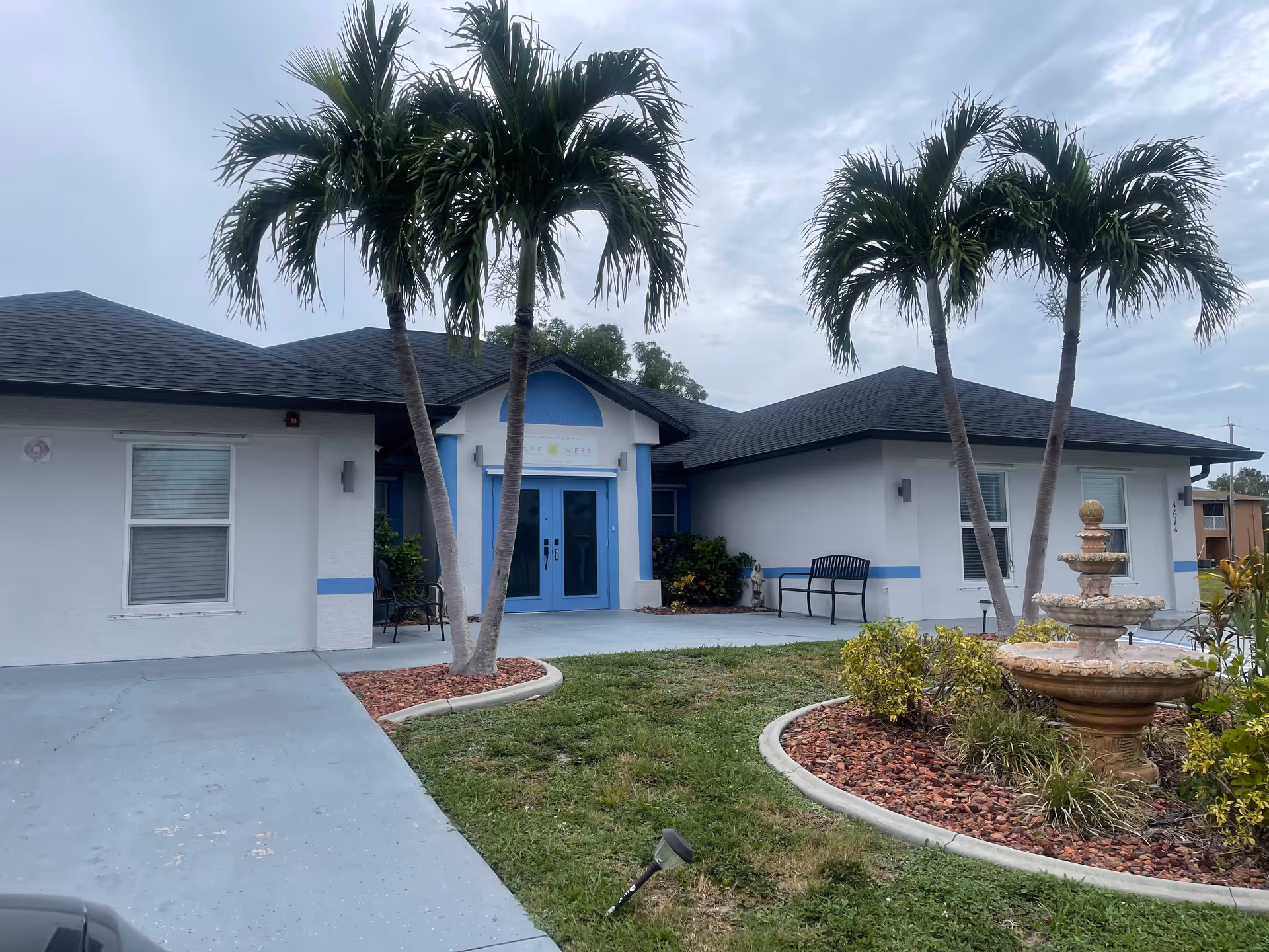 Single-story building exterior with blue double doors, palm trees, benches and a small fountain in the front yard.