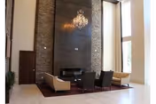 A spacious and modern living room area with high ceilings, a large stone fireplace, a chandelier, and tall windows letting in natural light. The seating arrangement includes two beige sofas and three dark chairs around a rectangular rug.