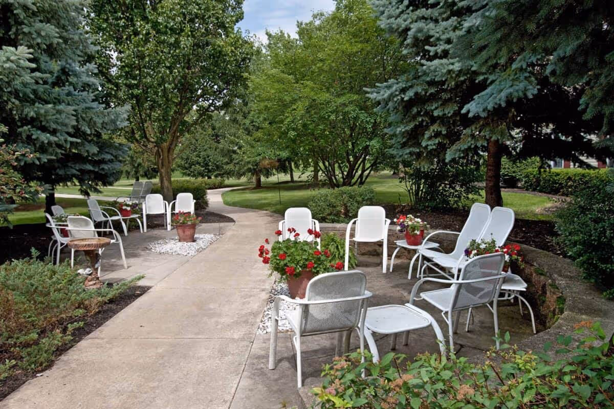 Outdoor seating area with white metal chairs and small tables arranged on a concrete patio surrounded by green trees, shrubs, and potted red flowers. A paved walkway curves through the landscaped garden area under a partly cloudy sky.