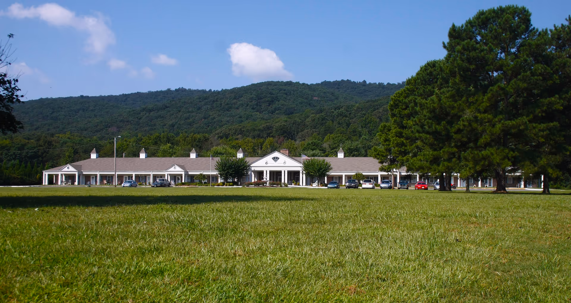 Single-story senior living facility building with cars parked in front, a wide grassy lawn in the foreground and wooded hills behind.