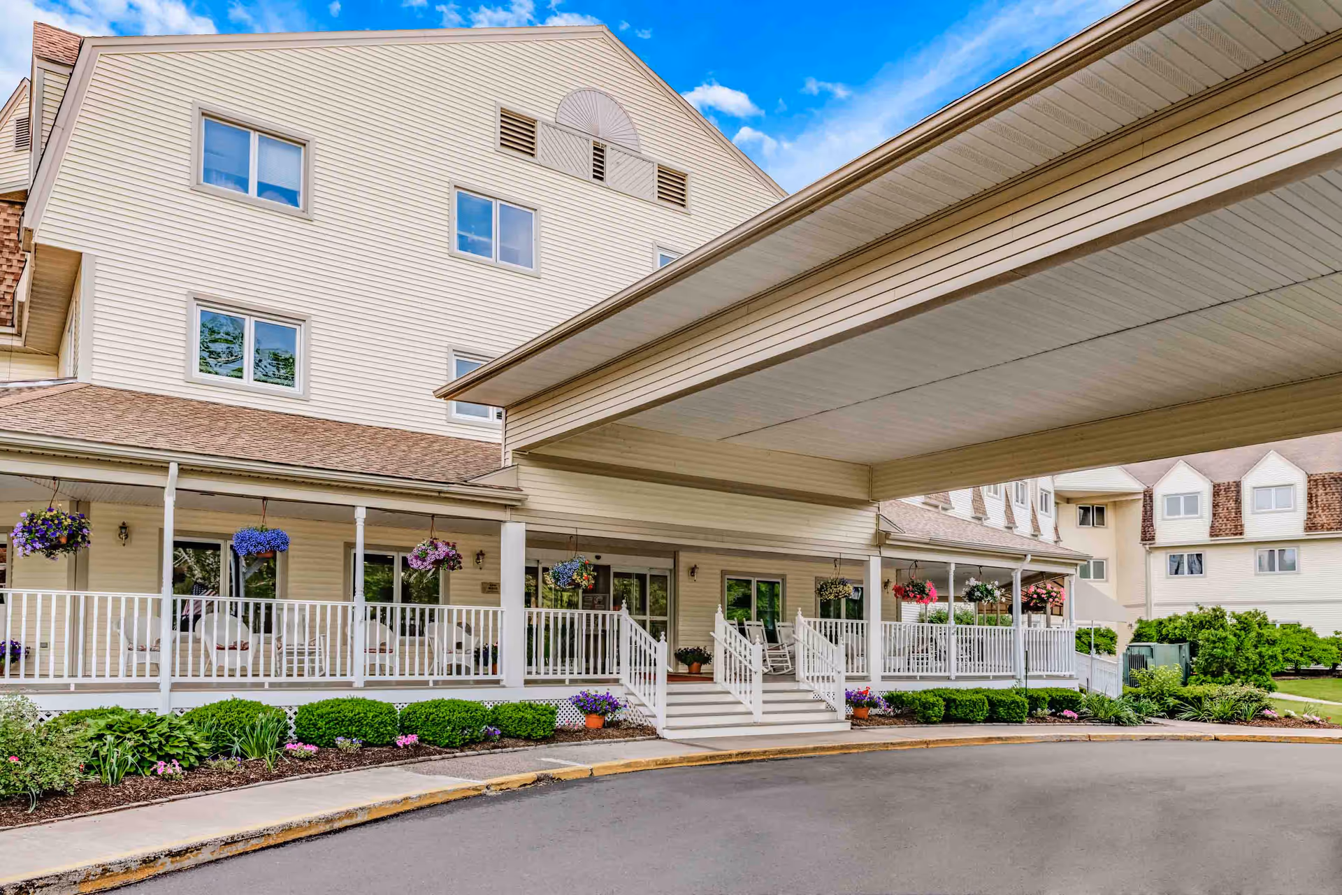 Exterior view of Holiday Farmington senior living facility showing a large covered entrance with white railings and steps leading to the front doors. The building has beige siding, multiple windows, and hanging flower baskets along the porch. There are well-maintained shrubs and flowers along the sidewalk and a clear blue sky with some clouds above.