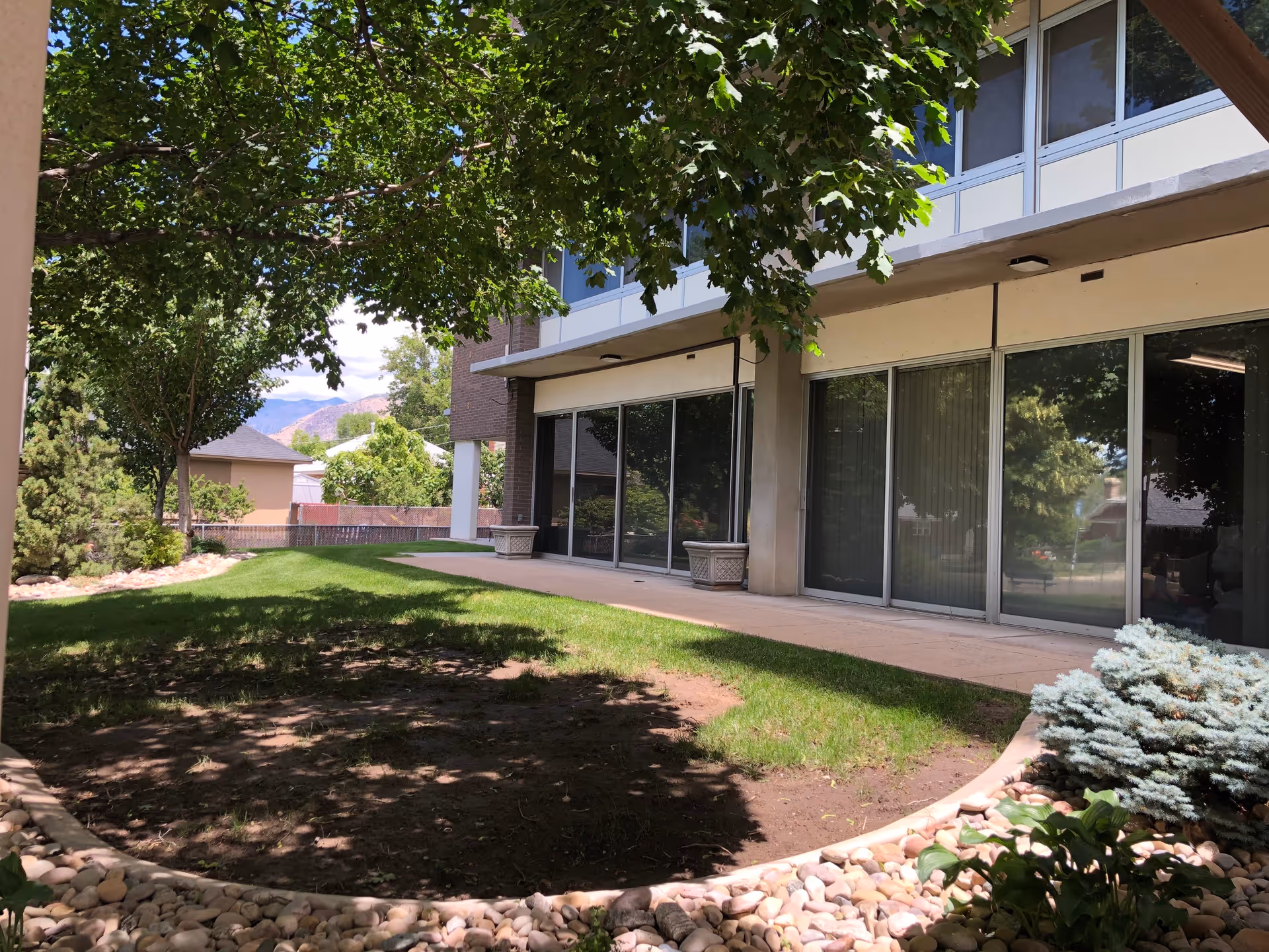 Outdoor view of a senior living facility with a grassy area, trees providing shade, a concrete walkway, and large windows on the building reflecting the surrounding greenery.