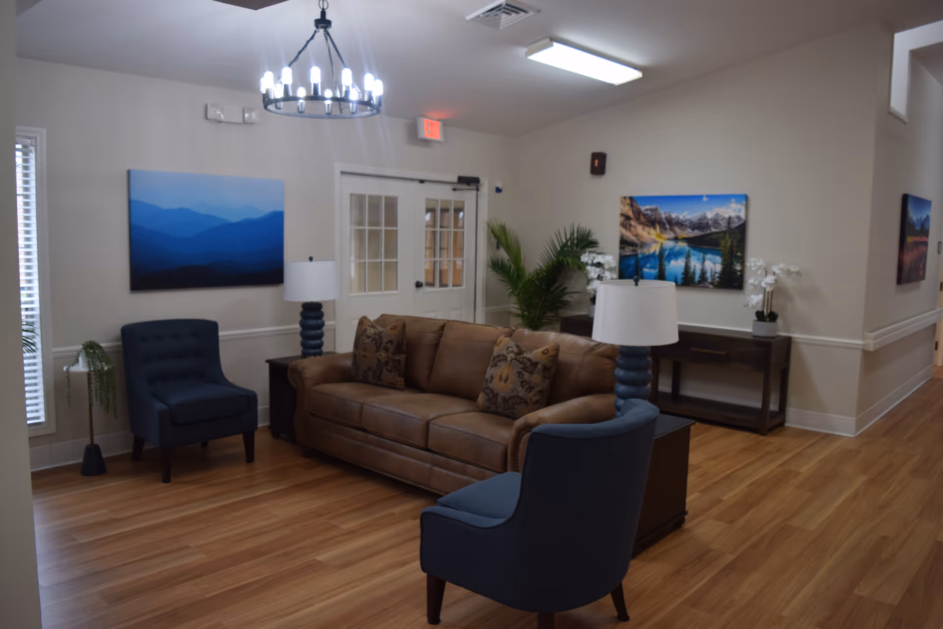 A furnished common lounge with a brown leather sofa, two blue armchairs, lamps, wall art, and wood flooring.