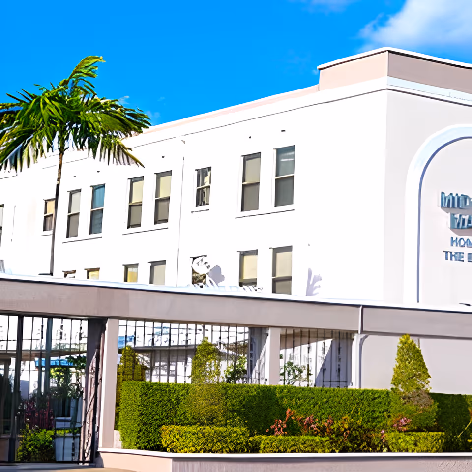 Exterior view of Midtown Manor, a multi-story white building with several windows, surrounded by neatly trimmed green hedges and a palm tree under a clear blue sky.