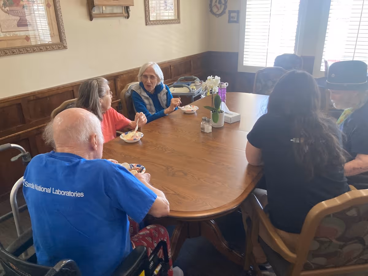 Five people sitting around a wooden dining table in a room with wood-paneled walls and framed artwork. They appear to be eating and conversing. One person is in a wheelchair wearing a blue shirt, and another person is wearing a black hat. There is a small white flower pot and a tissue box on the table.