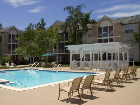 Outdoor swimming pool area with several lounge chairs arranged around the pool. There is a white pergola structure on one side and a large building with multiple windows and balconies in the background. Palm trees and other greenery surround the pool area under a clear blue sky.