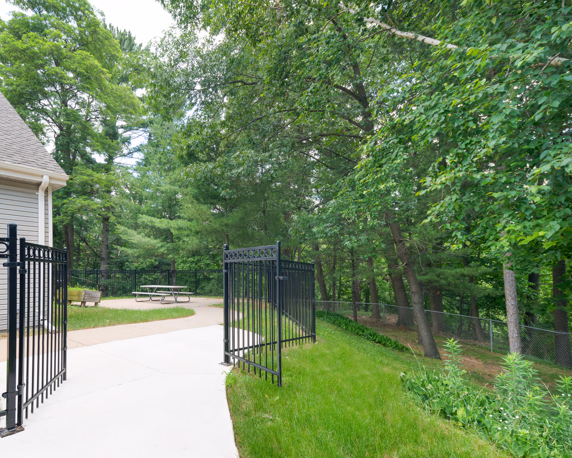 Outdoor area with a paved walkway leading through an open black metal gate. There is a picnic table on a concrete pad surrounded by grass and trees, with a building partially visible on the left side.