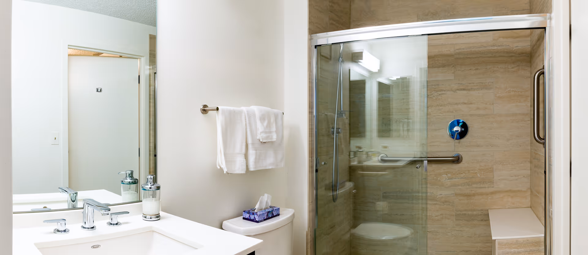 A clean and modern bathroom featuring a white sink with a chrome faucet, a soap dispenser, a large mirror, a towel rack with white towels, a toilet with a tissue box on top, and a glass-enclosed shower with beige tiled walls and a built-in bench.