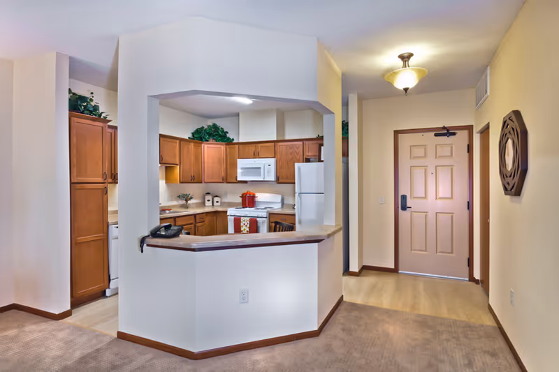 Open-plan kitchen with wooden cabinets, white appliances and a pass-through counter next to the apartment entry door.