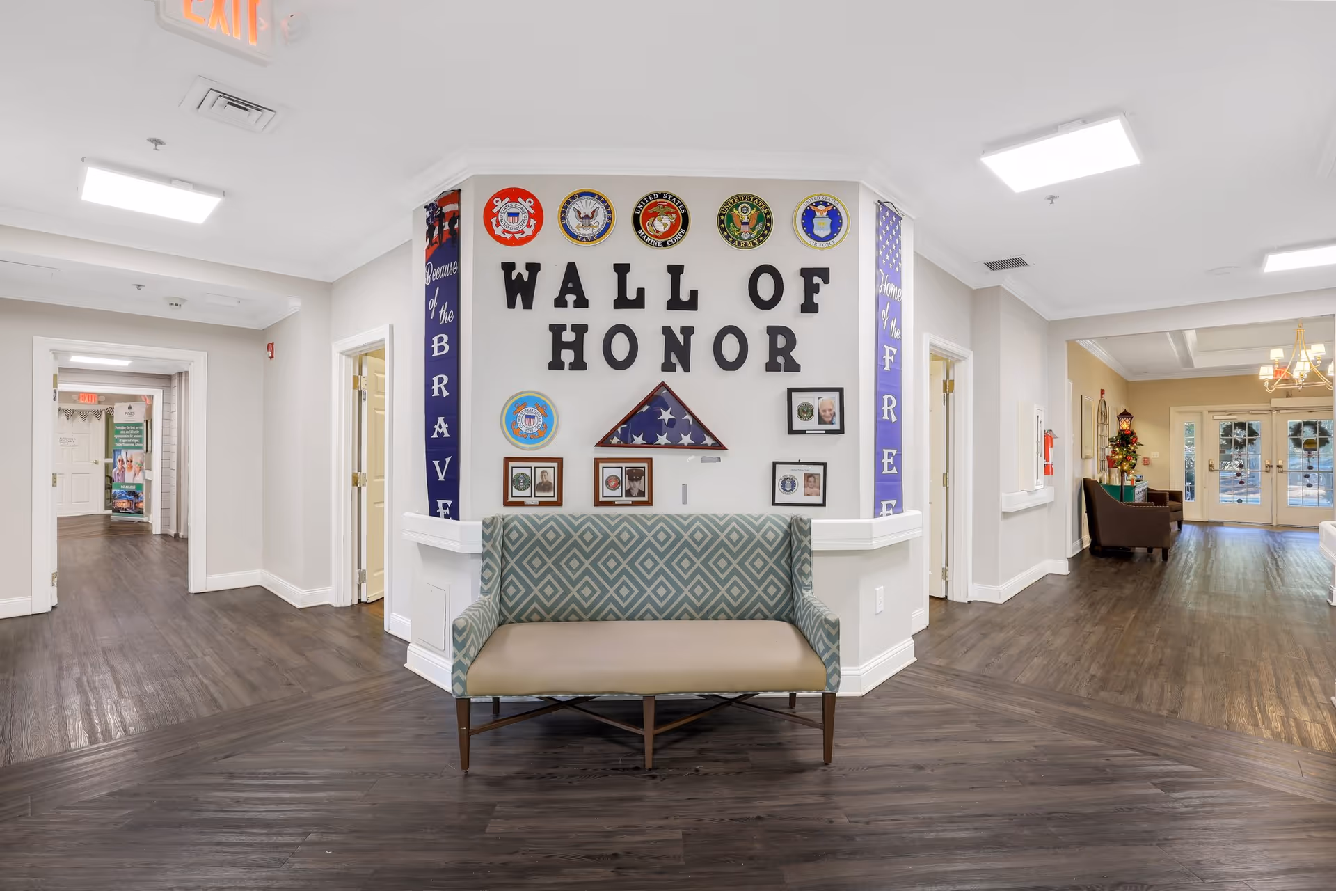 Interior hallway of The Pines at Bluffton Assisted Living & Memory Care Community featuring a 'Wall of Honor' display with military emblems, photos, and a folded American flag. A patterned bench is placed in front of the wall, and the hallway has wood flooring, white walls, and ceiling lights. Doors and exit signs are visible in the background.