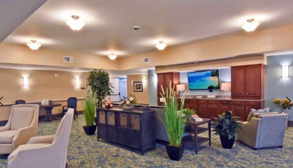 A spacious and well-lit common area in a senior living facility featuring comfortable armchairs, a large flat-screen TV mounted on a wall with wooden cabinetry below, several potted plants, and a carpeted floor with a floral pattern. The room has soft beige walls with wall sconces and ceiling lights providing warm illumination.