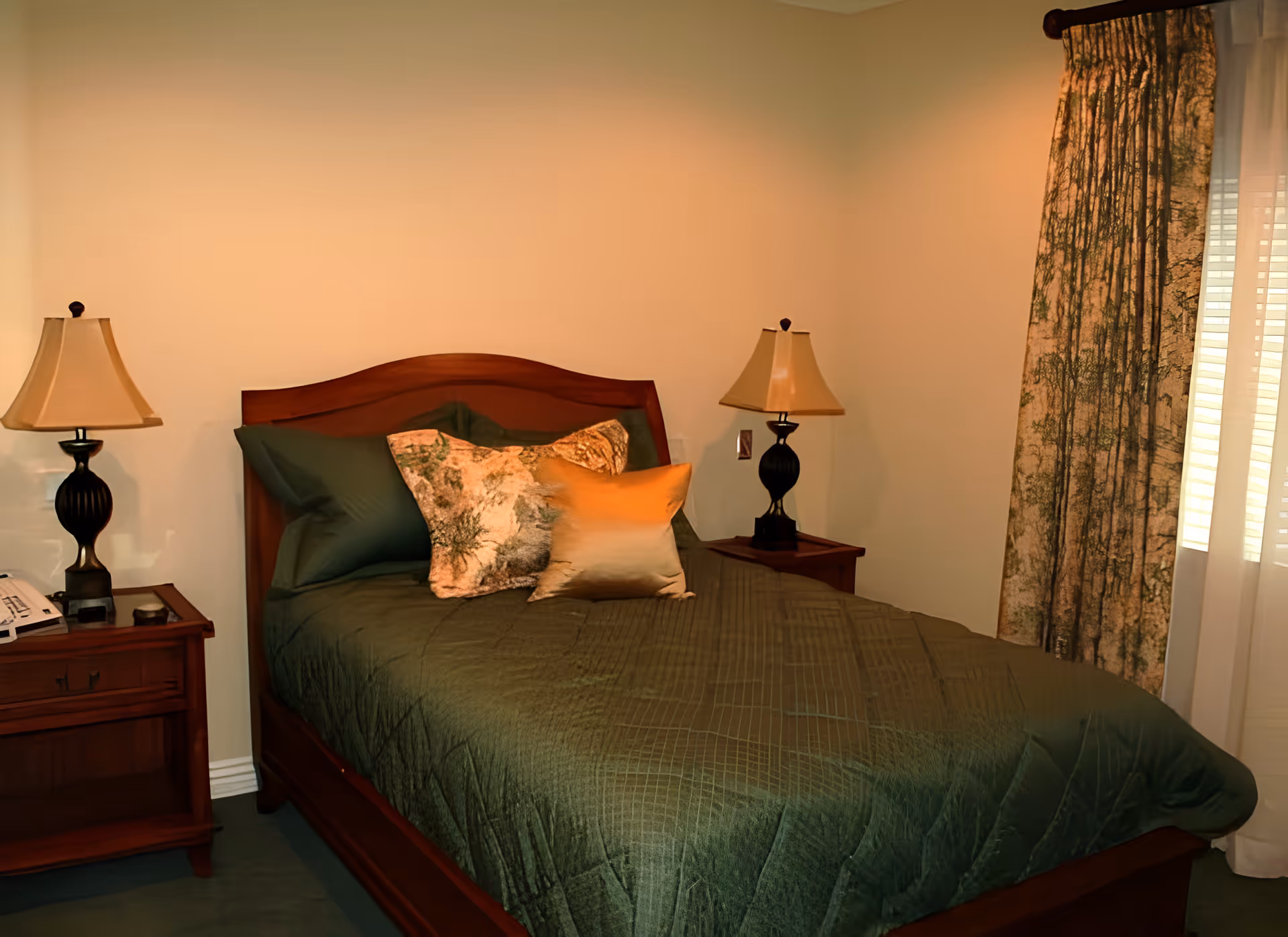 A cozy bedroom with a wooden bed frame, green bedding, and three decorative pillows. There are two matching bedside tables, each with a lamp. A window with patterned curtains is visible on the right side of the room.