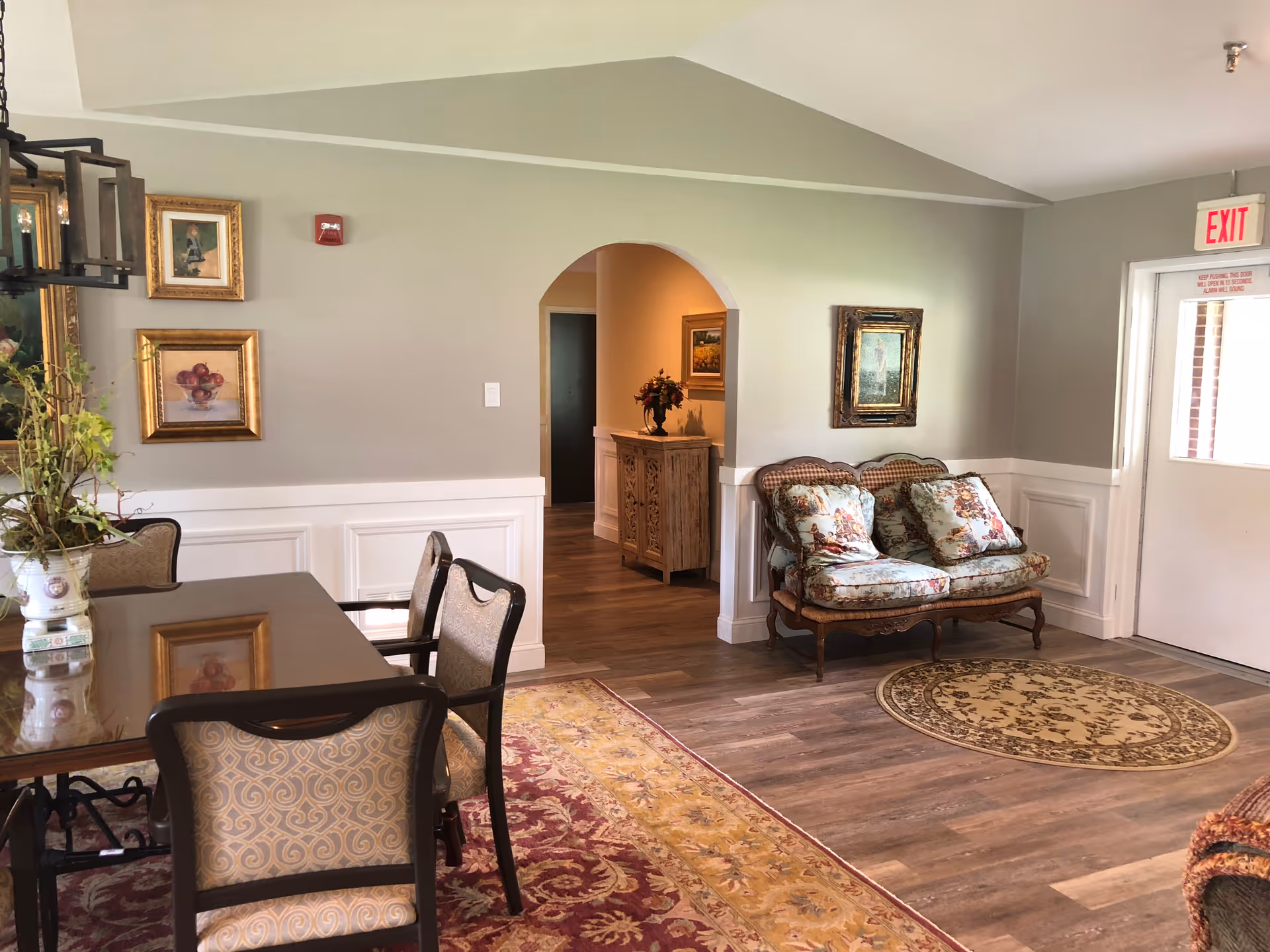 A cozy interior room with a wooden dining table and upholstered chairs on a patterned rug. There is a small vintage-style loveseat with floral cushions against the wall near an exit door. The walls are decorated with framed paintings and there is a wooden cabinet visible through an arched doorway. The flooring is wood with an additional round rug near the loveseat.