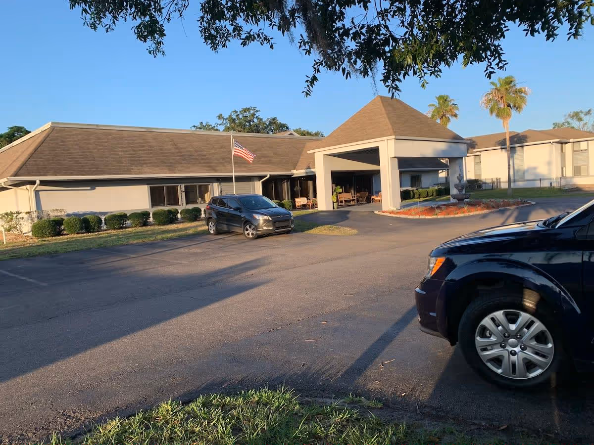 Exterior view of Oakview Terrace Assisted Living and Memory Care building with a covered entrance, an American flag on a pole, two parked cars, and palm trees in the background under a clear blue sky.