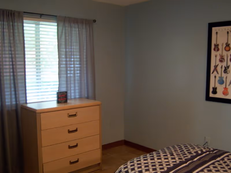 Small bedroom with a wooden dresser beneath a curtained window, a corner of a bed visible and a framed guitar print on the wall.