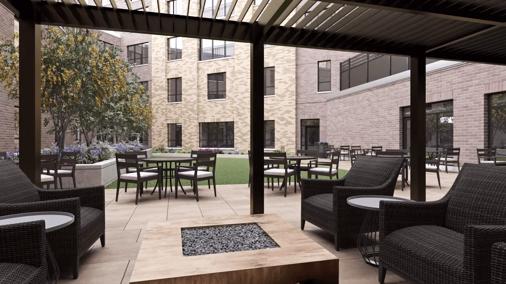 Outdoor courtyard area with a covered seating space featuring black wicker chairs and small round tables. Beyond the covered area, there are multiple tables and chairs on a tiled patio surrounding a grassy lawn with landscaping and trees. The courtyard is enclosed by a multi-story brick building with numerous windows.