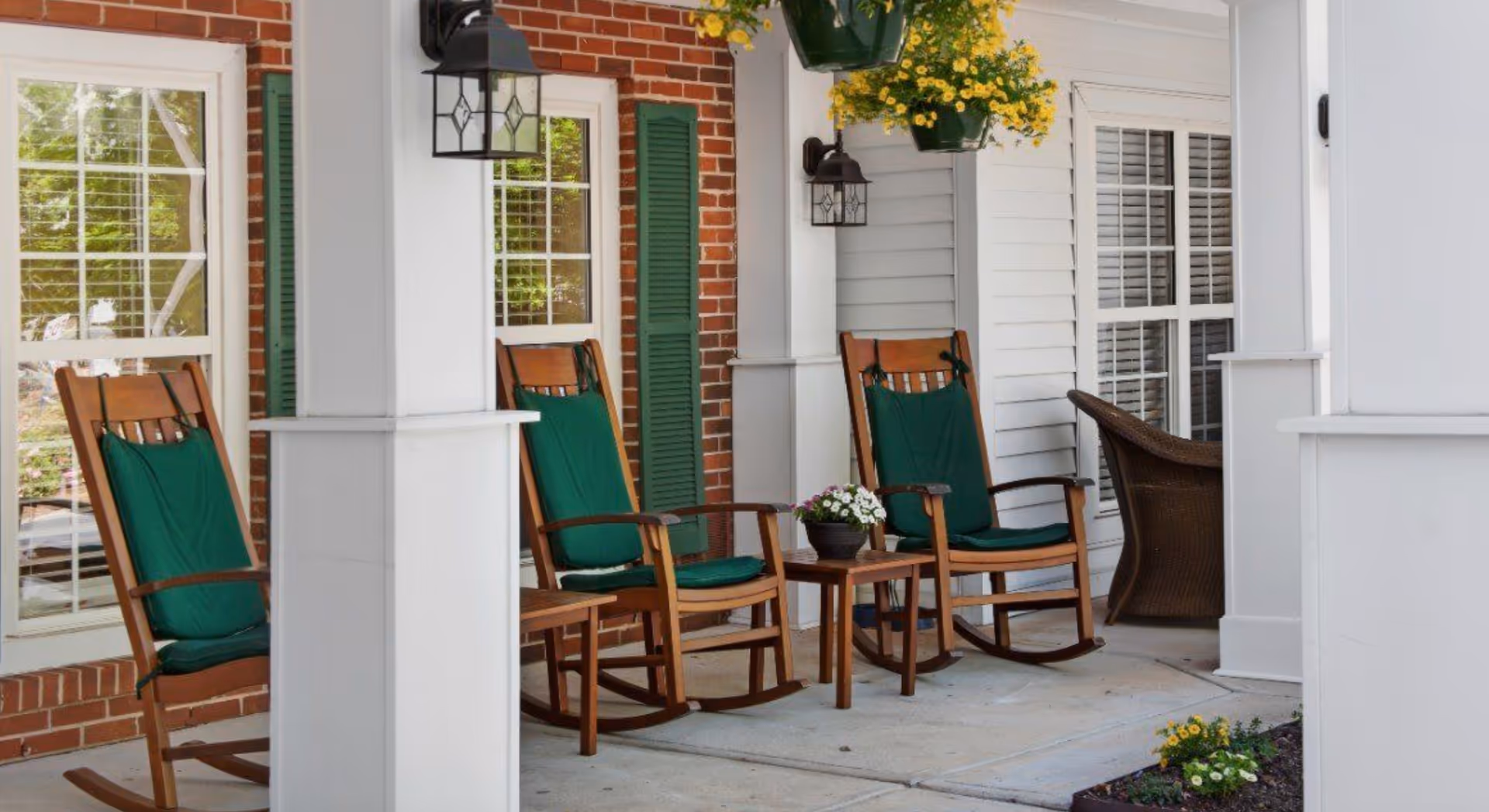 Covered front porch with wooden rocking chairs with green cushions, a small table with potted flowers, and hanging planters at a brick-and-siding building.