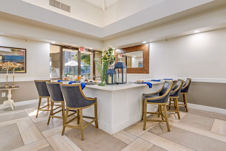 A bright dining area with a large white rectangular table surrounded by eight wooden chairs with black cushions. The table is set with plates, glasses, and blue napkins. There are decorative lanterns and a flower arrangement in the center of the table. A large mirror hangs on the wall behind the table, and a glass door with an exit sign leads outside where an umbrella and greenery are visible.