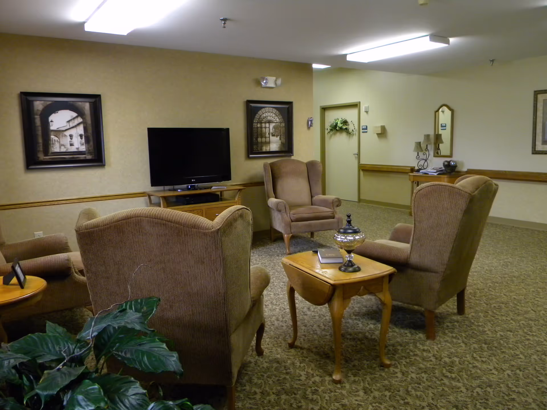 A cozy common area with four upholstered armchairs arranged around a wooden side table holding a decorative item and a book. A flat-screen TV is mounted on a wooden stand against the wall, which is decorated with framed artwork. The room has beige walls, carpeted flooring, and fluorescent ceiling lights. There is a door with a wreath and a hallway visible in the background.