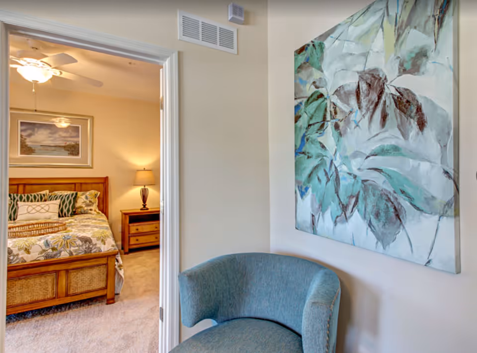 View of a cozy bedroom with a wooden bed frame, floral bedding, a nightstand with a lamp, and a ceiling fan, seen through an open doorway from an adjacent room with a blue upholstered chair and a large abstract painting on the wall.