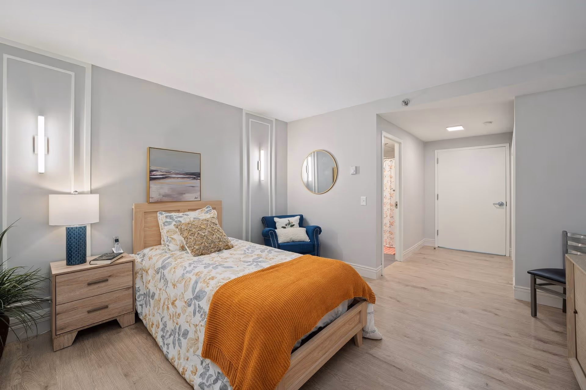 A bright and modern bedroom in a senior living facility featuring a single bed with floral bedding and an orange throw blanket. Next to the bed is a wooden nightstand with a blue lamp and a phone. A blue armchair with a pillow is placed against the wall, which has a round mirror and a framed landscape painting. The room has light wood flooring and neutral-colored walls, with a doorway leading to a bathroom and a closed white door in the background.