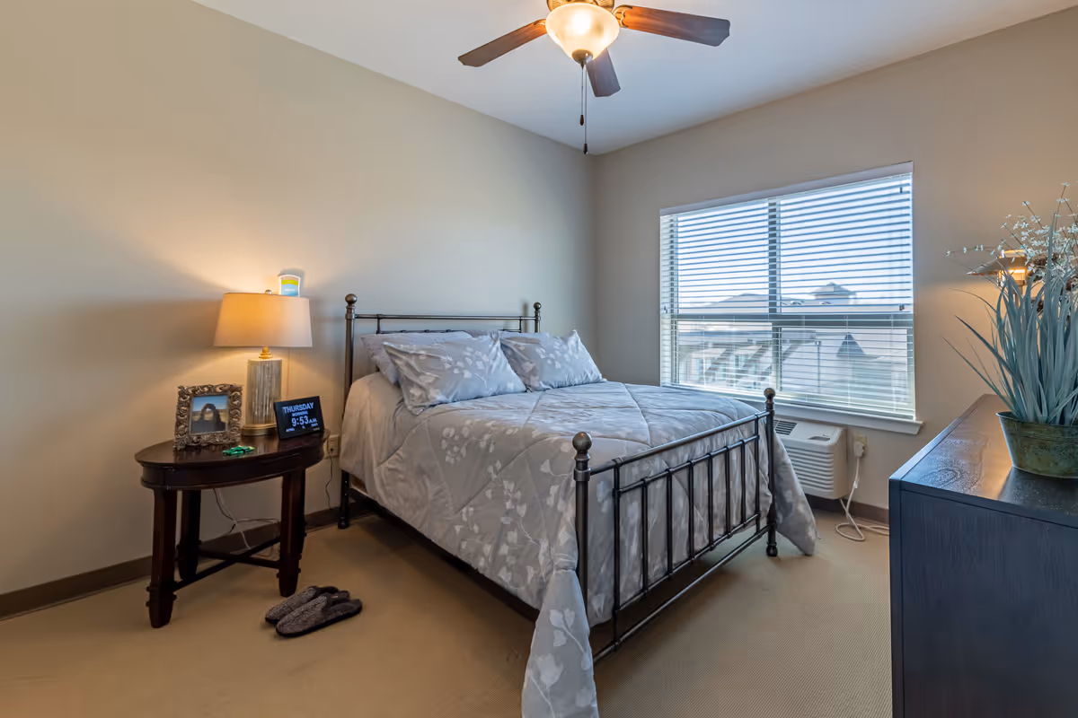 A tidy bedroom featuring a metal-framed bed with patterned bedding, a nightstand with a lamp and digital clock, a dresser, and a window with blinds.