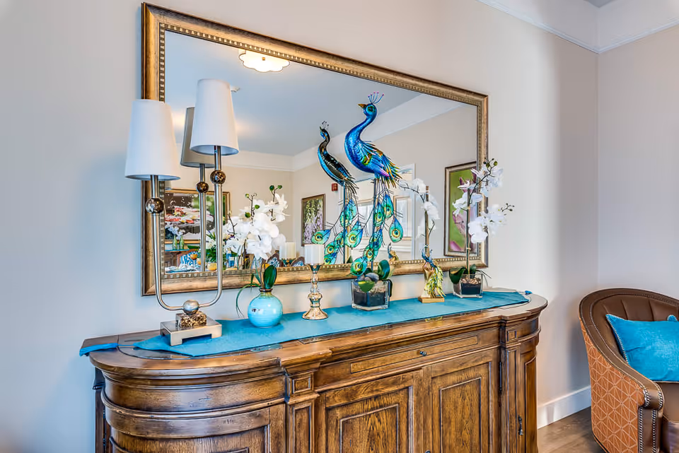 A wooden sideboard with a blue runner on top, decorated with two white orchid plants in vases, two colorful peacock figurines, and a modern lamp with two white shades. A large framed mirror hangs on the wall behind the sideboard, reflecting part of the room including artwork and lighting fixtures. To the right, there is a brown upholstered chair with a blue cushion.