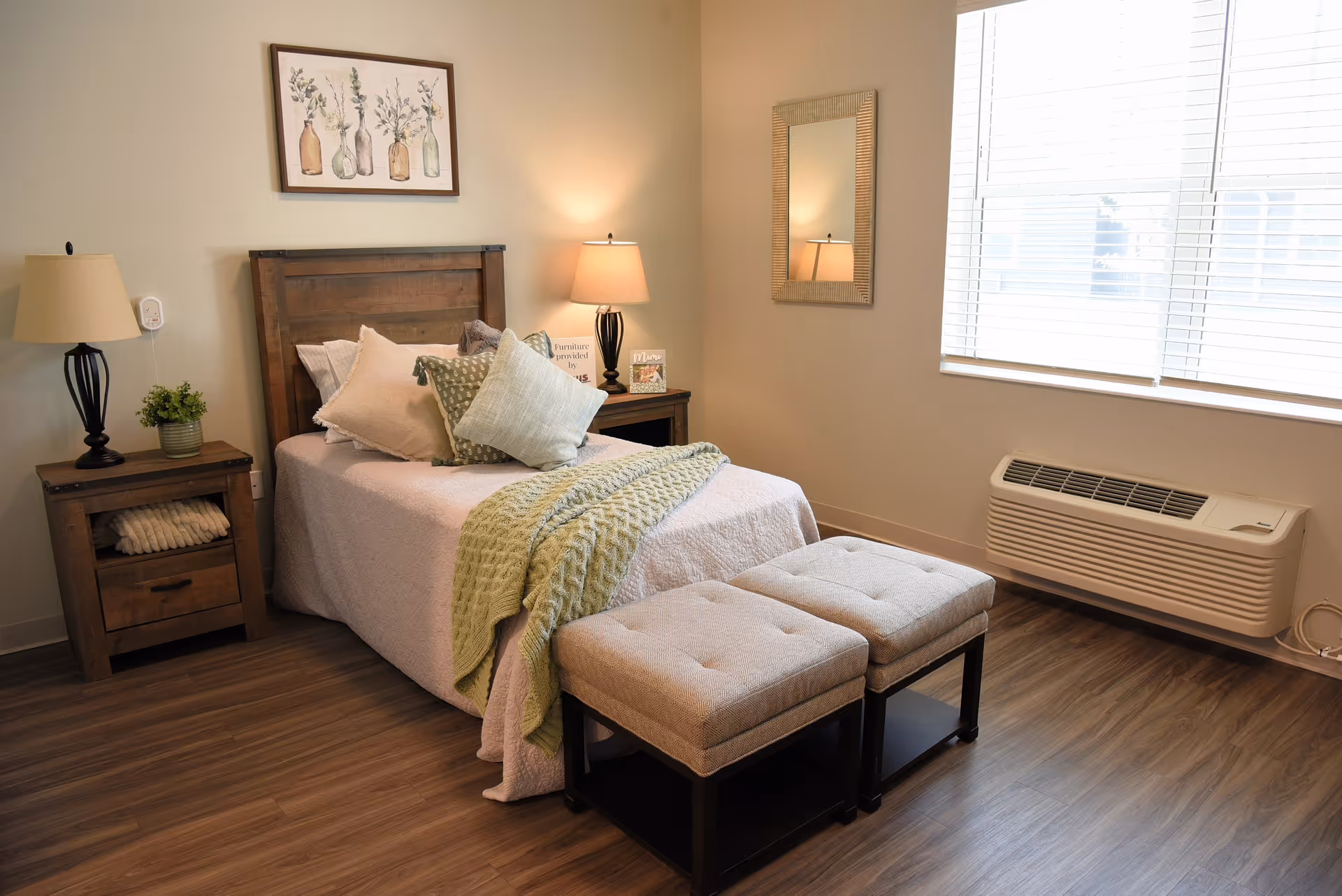 A cozy bedroom with a wooden bed frame, a neatly made bed with multiple pillows and a green knitted throw blanket. There are two cushioned stools at the foot of the bed, a wooden nightstand with a lamp and a small plant, a framed botanical artwork on the wall, a mirror on the adjacent wall, and a large window with blinds letting in natural light. The room has wood flooring and a wall-mounted air conditioning unit below the window.