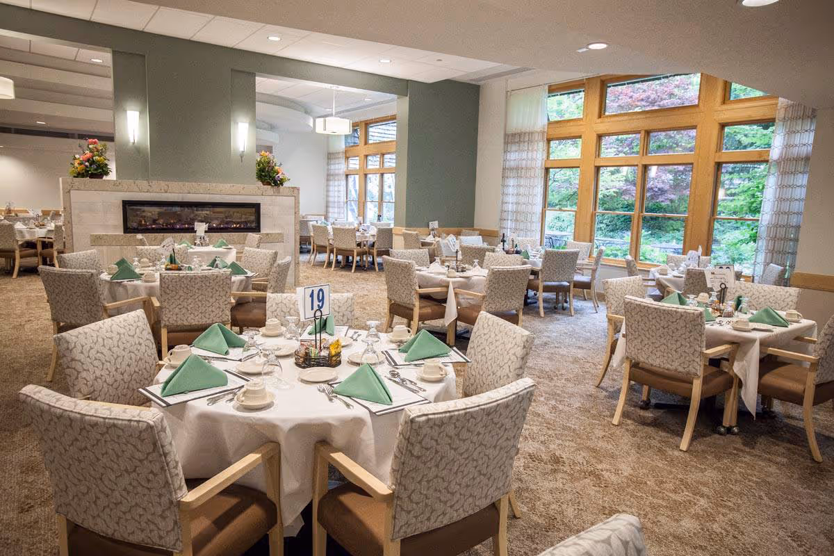 A spacious dining room in a senior living facility with multiple round tables set with white tablecloths, green folded napkins, cups, and silverware. The room features large windows with wooden frames allowing natural light and views of greenery outside. There is a modern fireplace in the center with floral arrangements on top, and comfortable upholstered chairs around each table.