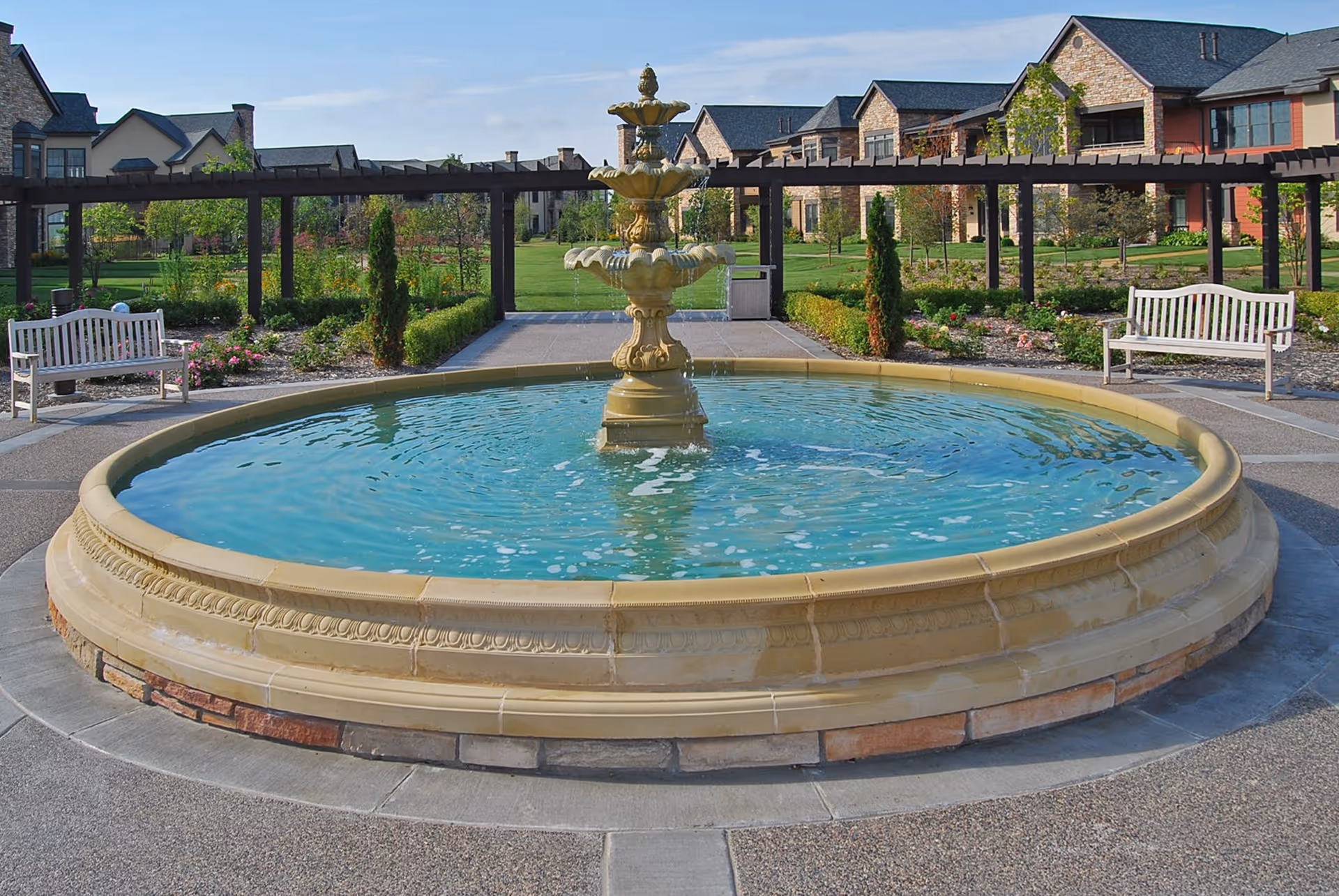 A large circular stone fountain with three tiers, filled with water, situated in an outdoor garden area. Surrounding the fountain are paved walkways, benches, and landscaped greenery with flowers and small trees. In the background, there are residential-style buildings with stone and brick facades under a clear blue sky.