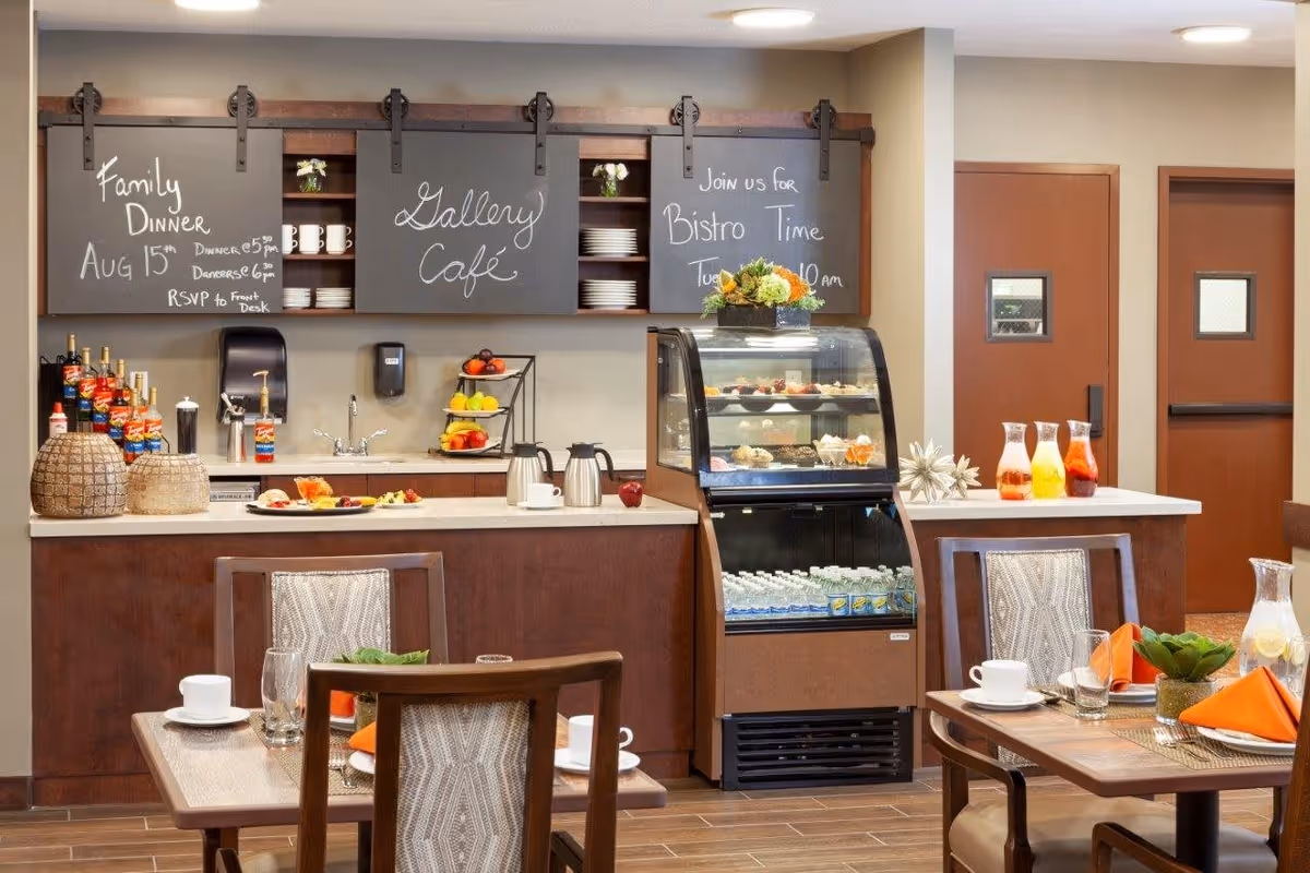 Interior view of a café area in a senior living facility with wooden tables and chairs set with cups, glasses, and orange napkins. A counter with a display case containing desserts and bottled drinks is visible, along with shelves holding plates, cups, and condiments. Chalkboard signs above the counter advertise a Family Dinner on August 15th and Bistro Time on Tuesday at 10 am.