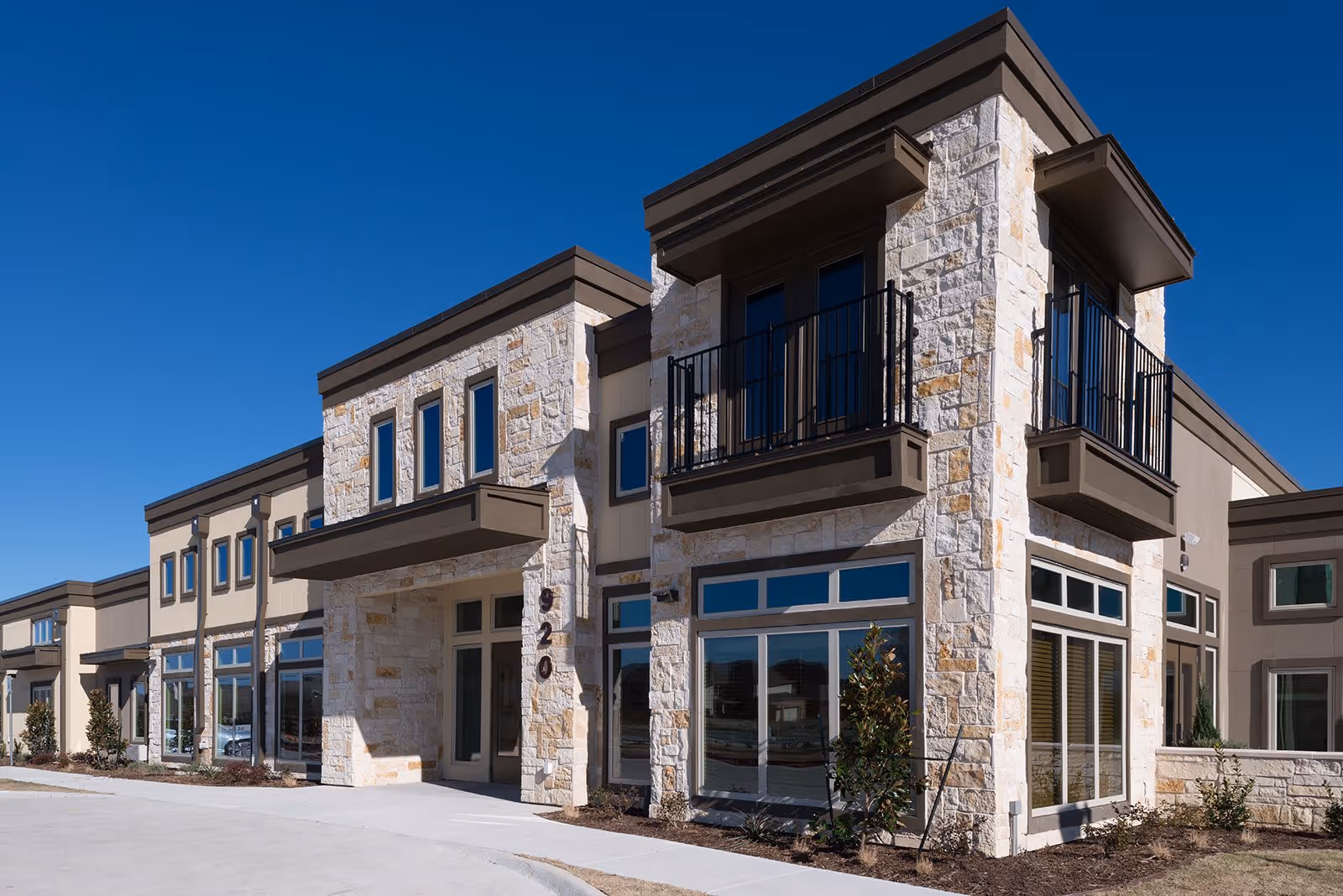 Exterior view of a modern two-story building with stone and beige stucco facade, large windows, and small balconies under a clear blue sky.