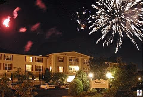 Nighttime view of Somerset Retirement Home and Assisted Living in Longview, WA, with fireworks lighting up the sky above the building. The building is illuminated with exterior lights and there are cars parked in front. A sign with the name Somerset is visible near the entrance.