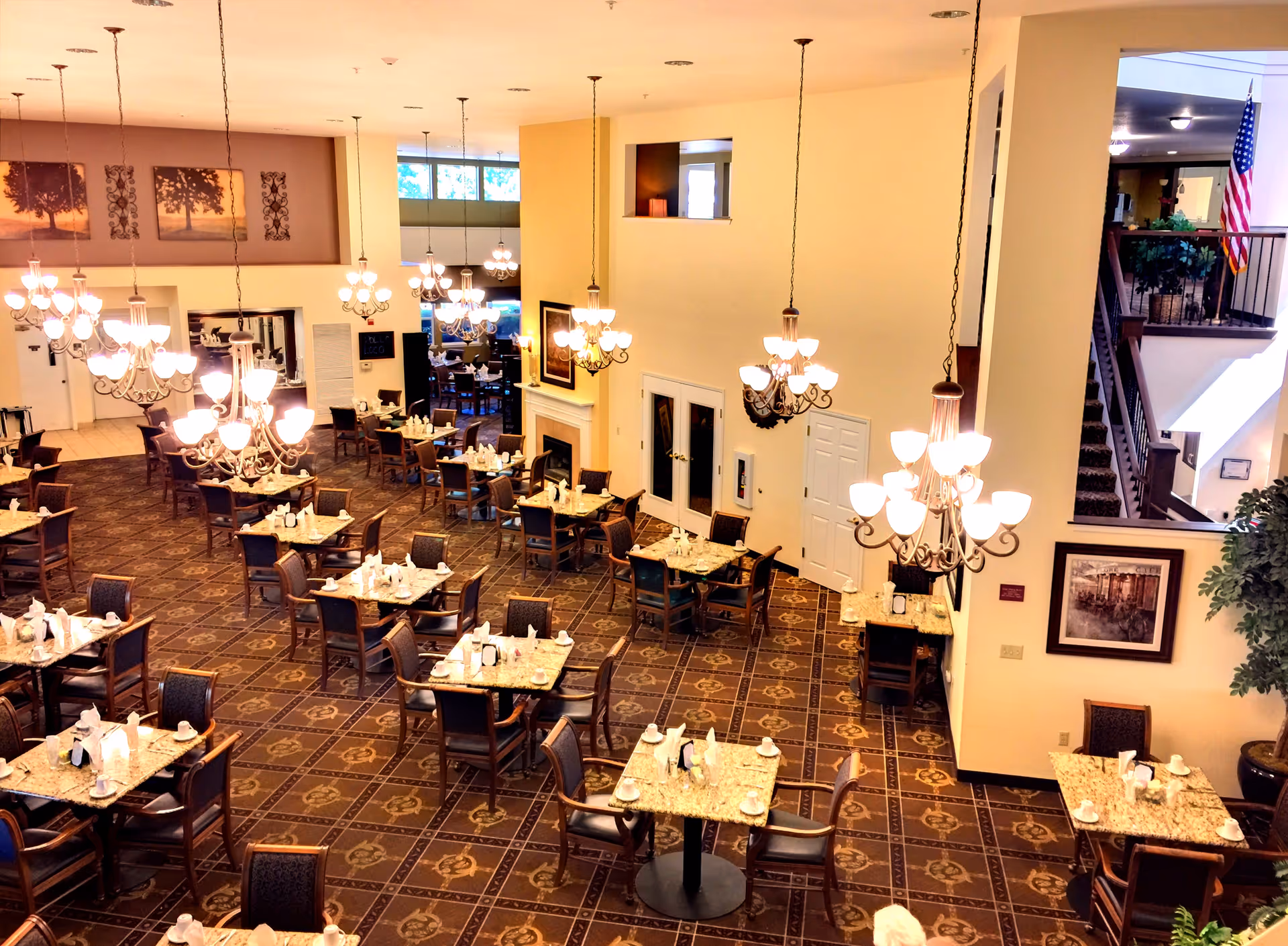 A spacious dining room with multiple tables set with cups, napkins, and utensils. The room features elegant chandeliers hanging from the ceiling, patterned carpet flooring, and a fireplace along one wall. There are framed pictures on the walls, large windows letting in natural light, and an American flag visible near a staircase in the background.