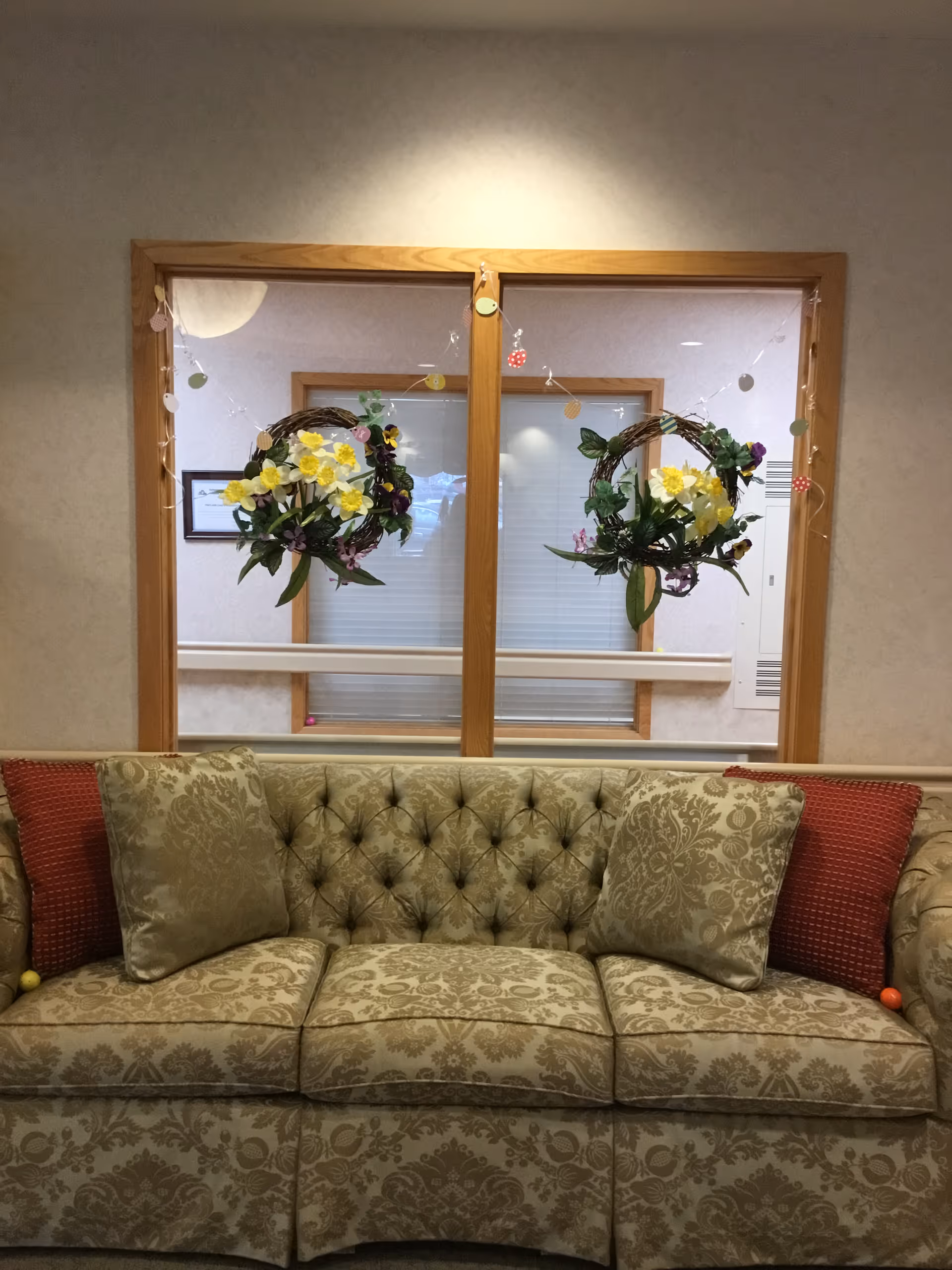 Tufted patterned sofa with cushions positioned beneath a window decorated with two hanging floral wreaths.