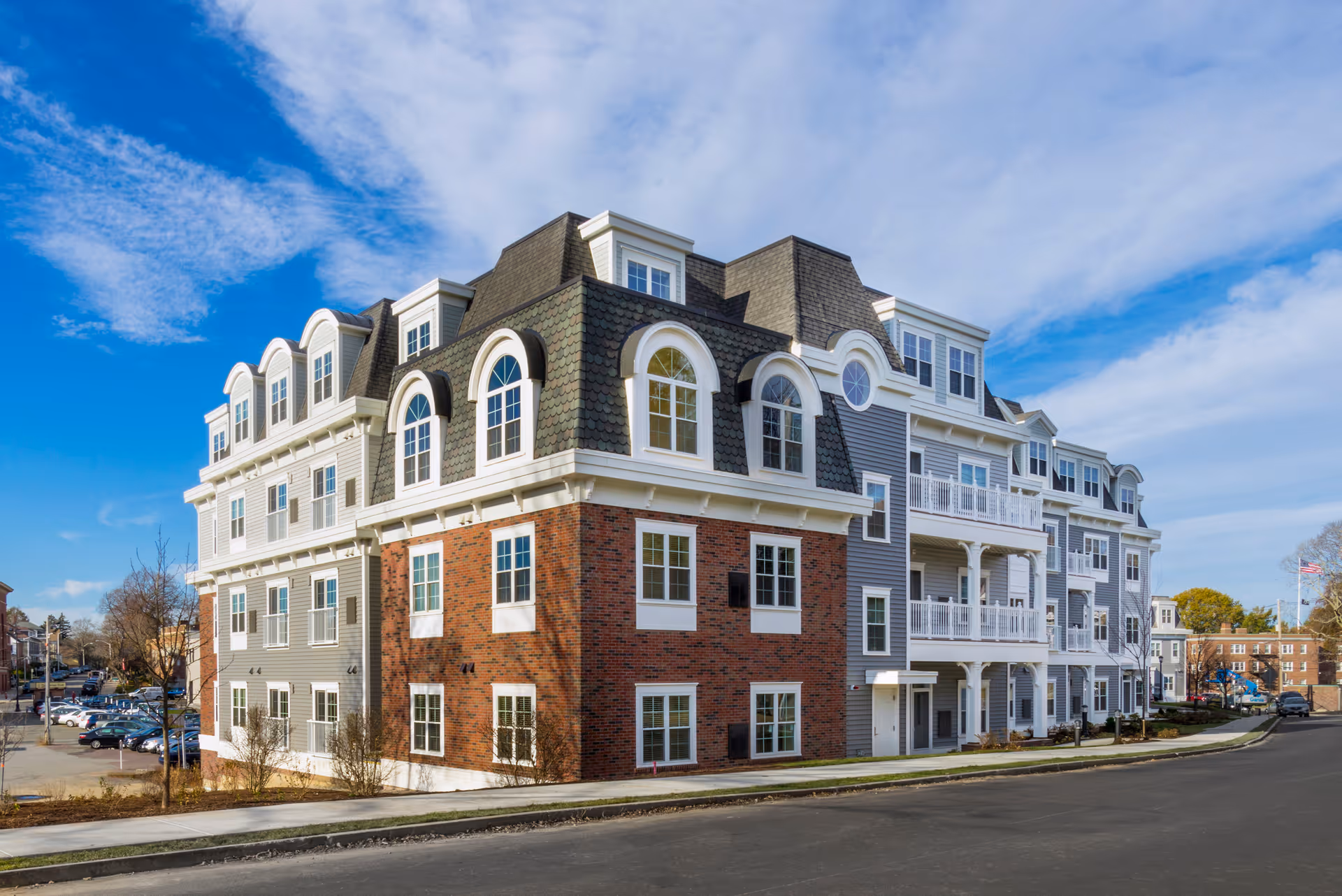 Multi-story senior living building with brick and gray siding, arched dormer windows and balconies under a blue sky.
