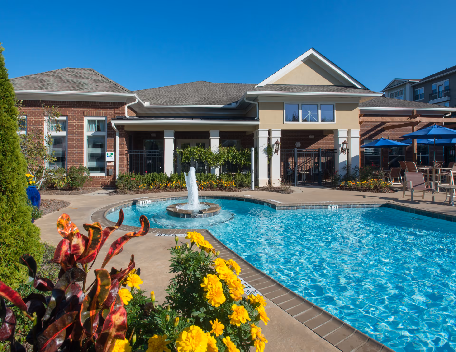 Outdoor swimming pool with a small fountain in the center, surrounded by a concrete deck with yellow flowers and plants in the foreground. Behind the pool is a brick building with white columns and a patio area with blue umbrellas and chairs under a clear blue sky.