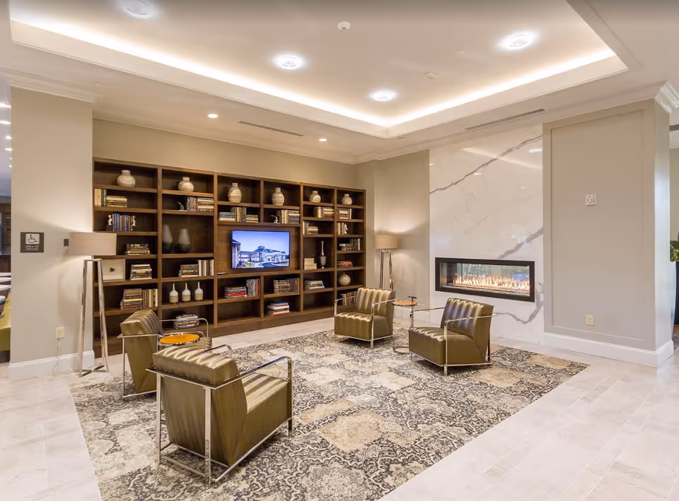 A modern living room area with four brown leather armchairs arranged around a patterned area rug. There is a long, rectangular gas fireplace set into a white marble wall on the right. On the left wall, there is a large built-in wooden bookshelf filled with books and decorative vases, with a TV screen displaying an image of a building. Two floor lamps with beige shades are positioned near the chairs. The ceiling has recessed lighting and a tray ceiling design.