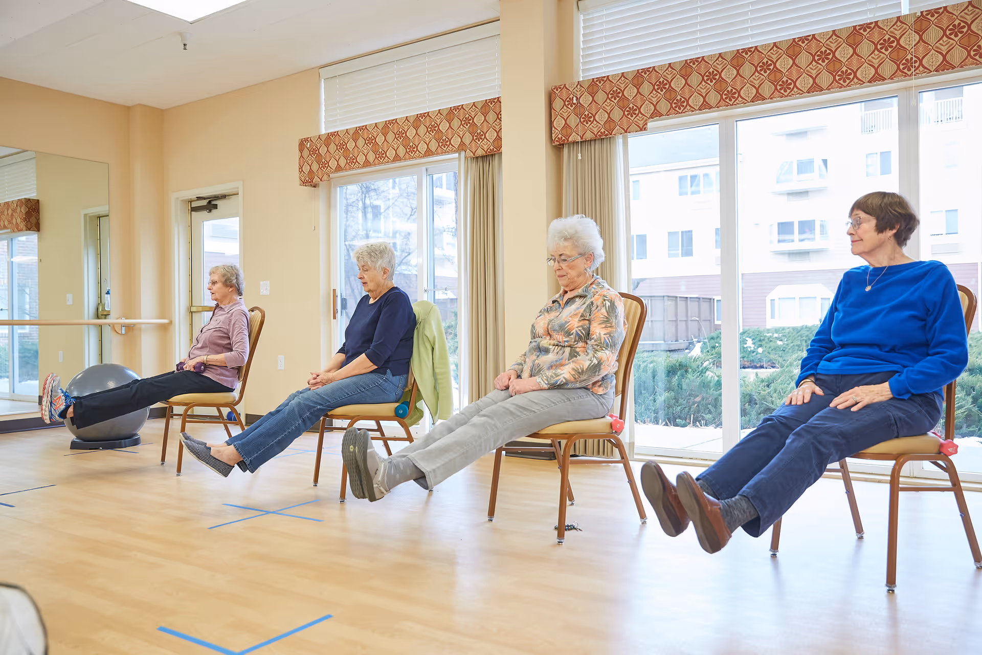 Four elderly women seated on chairs in a bright room with large windows, participating in a seated leg exercise, lifting their legs in unison. The room has light-colored walls, wooden flooring, and a large mirror on one side.