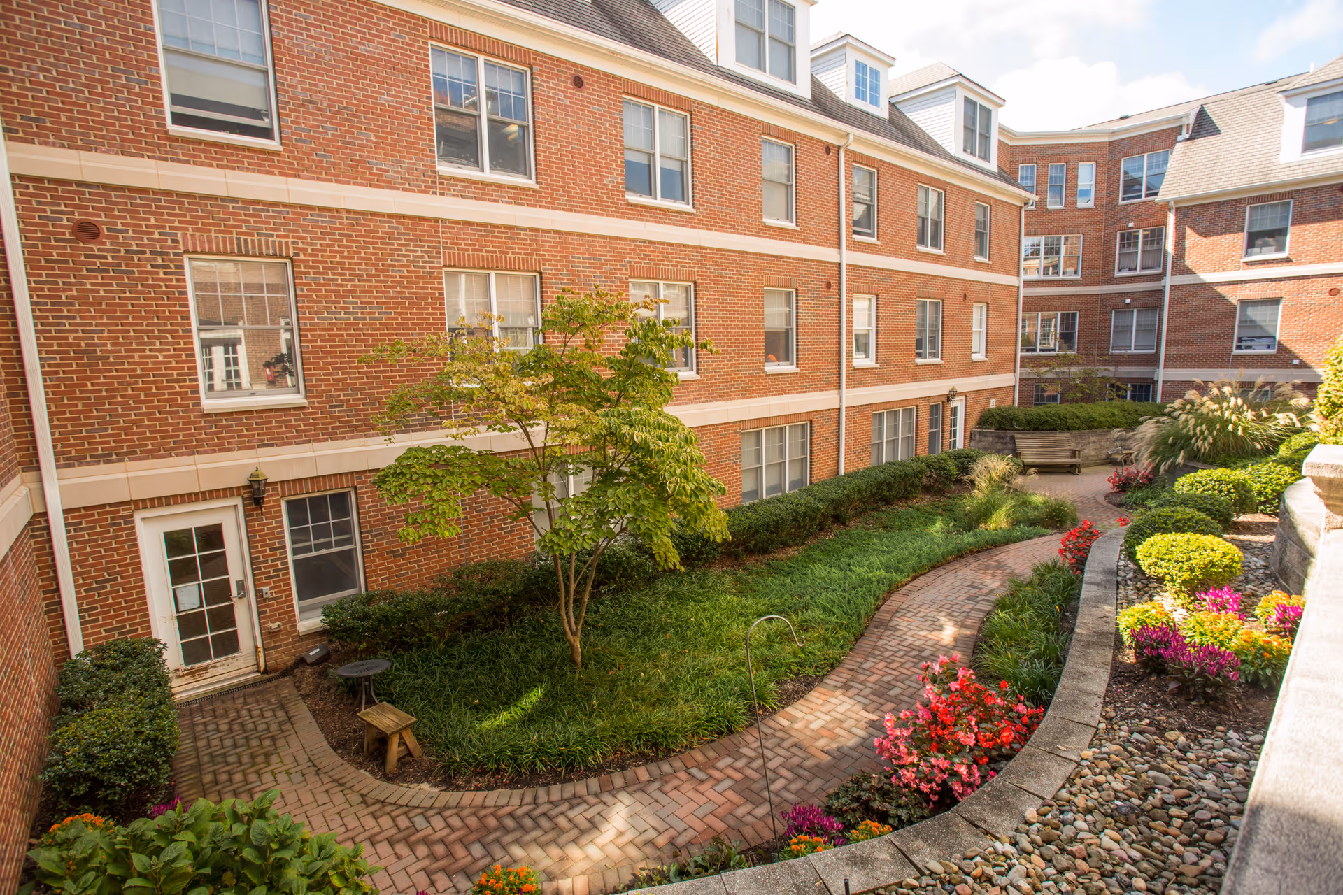 A landscaped courtyard area within a senior living facility featuring a curved brick pathway, green shrubs, colorful flowers, a small tree, and benches. The courtyard is surrounded by a multi-story red brick building with multiple windows.