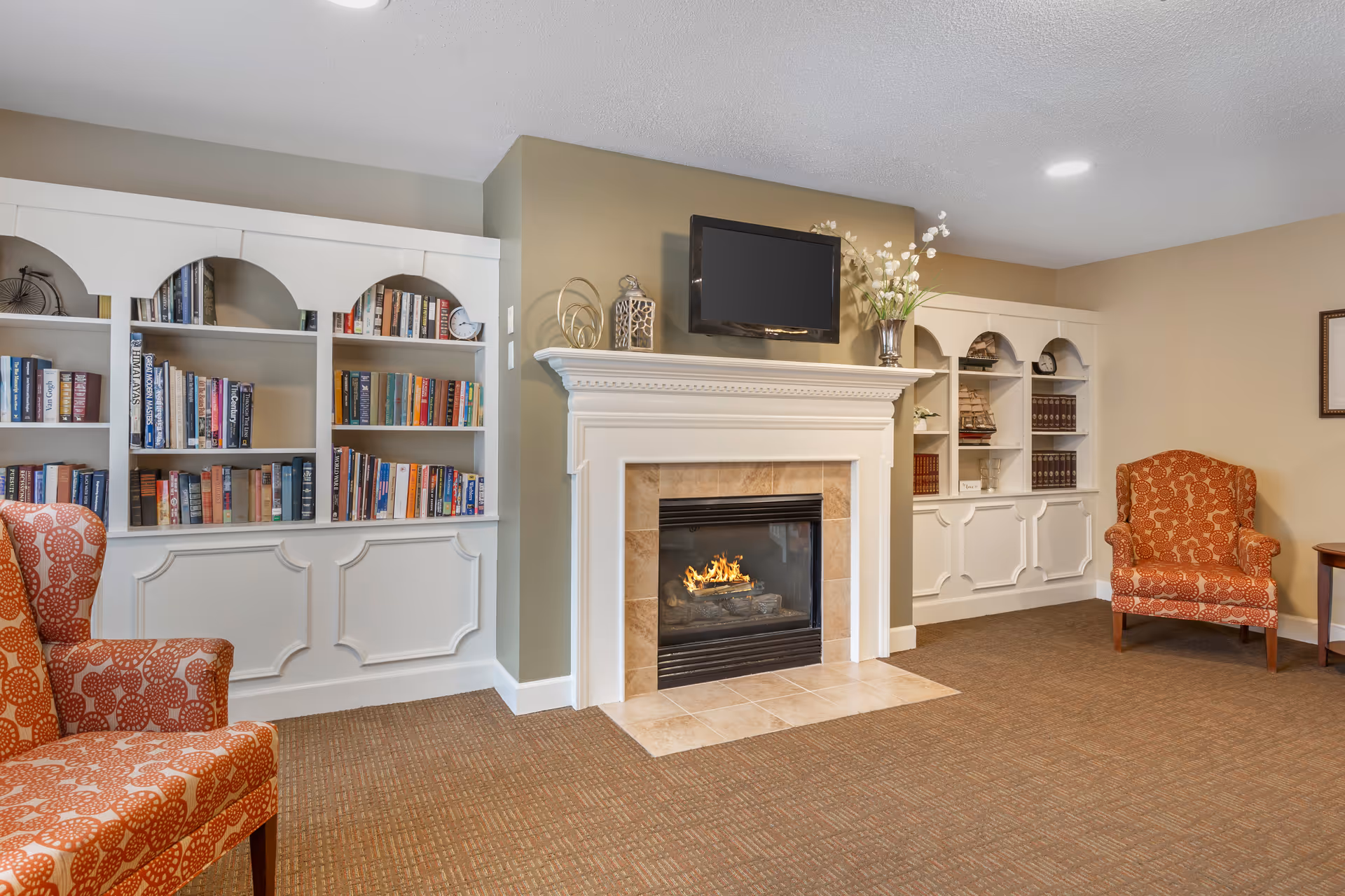 Cozy sitting room with a fireplace, wall-mounted TV, built-in white bookshelves, and patterned armchairs.