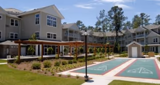 Outdoor view of Summerville Estates senior living facility showing a landscaped courtyard with shuffleboard courts, a wooden pergola, lamp posts, and multi-story residential buildings in the background surrounded by trees.