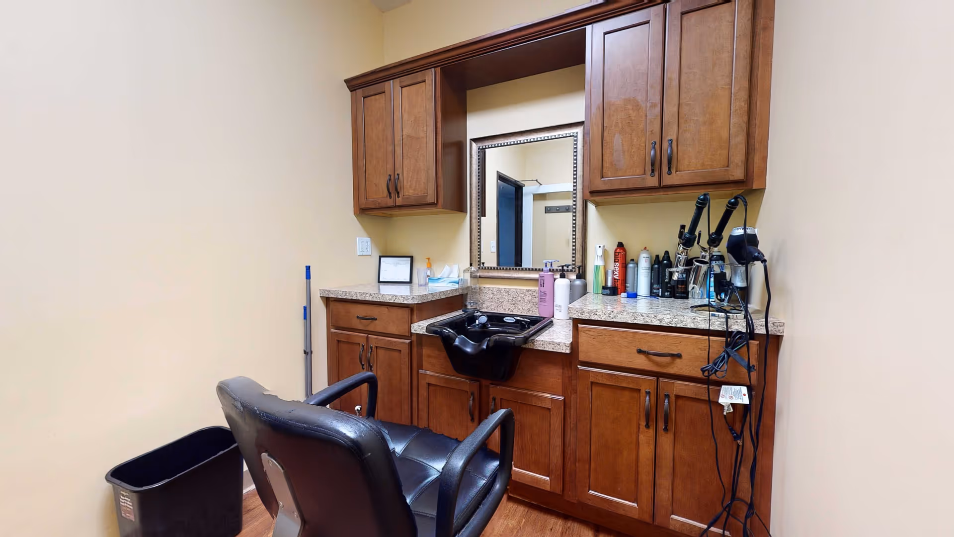 A small hair salon area with a black salon chair facing a countertop with a built-in black sink. Above the countertop are wooden cabinets and a mirror. Various hair care products and styling tools, including hair dryers and curling irons, are placed on the countertop. The walls are painted beige, and there is a black trash bin and a mop in the corner.