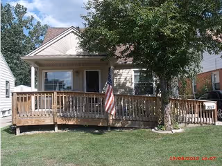 Front exterior view of a single-story house with a wooden ramp and railing leading to the entrance. There is an American flag mounted on the railing and a tree in the front yard. The house has light-colored siding and a small porch area.