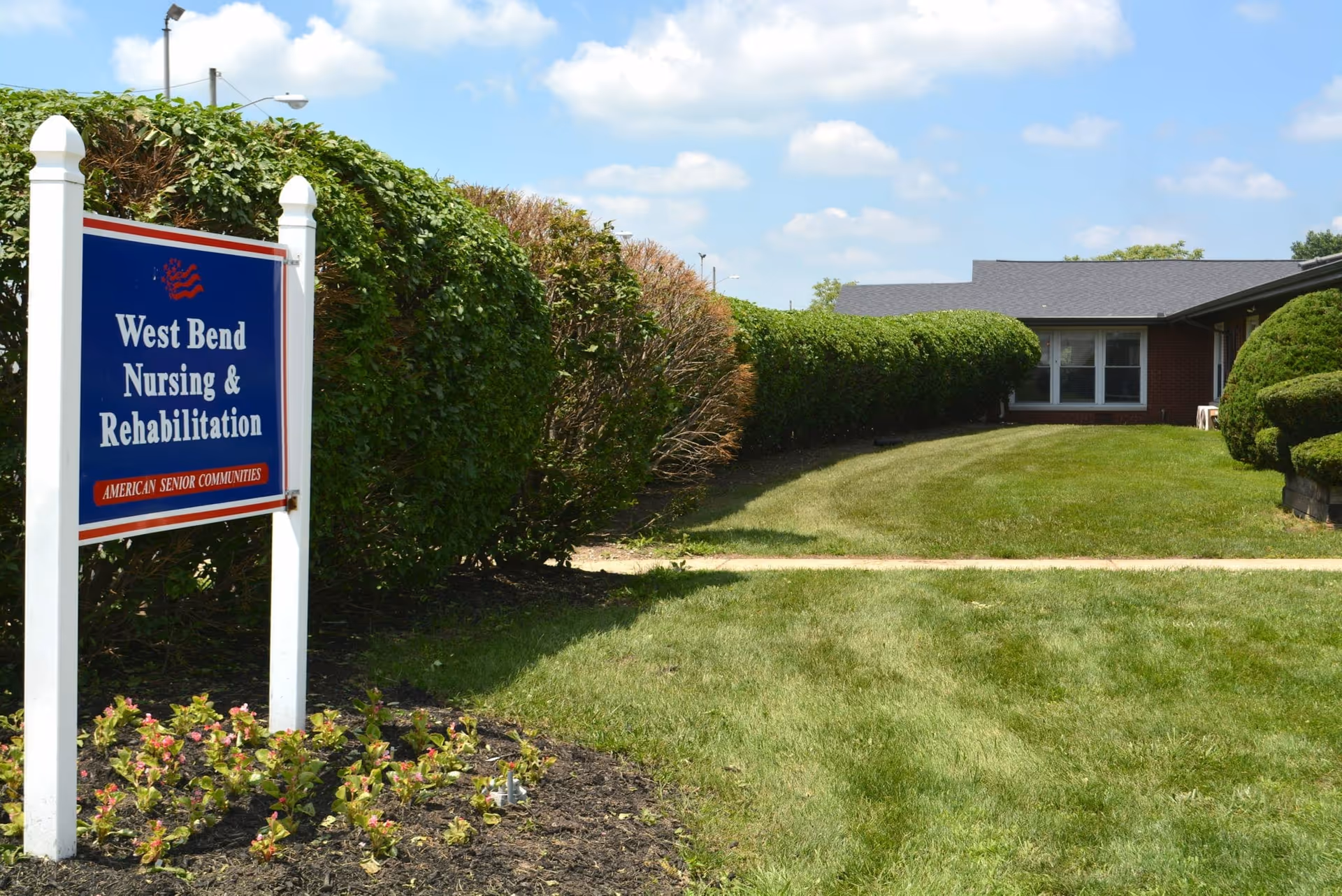 Outdoor view of West Bend Nursing & Rehabilitation facility with a sign in the foreground and a building with windows in the background, surrounded by neatly trimmed bushes and green grass under a blue sky with some clouds.