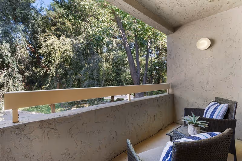 A small outdoor balcony area with two wicker chairs featuring blue and white striped cushions, a glass-top side table with a small potted plant, and a view of green trees beyond the balcony railing.